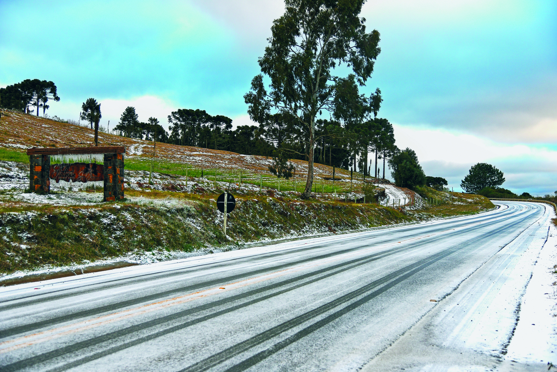 Fotografia. Rodovia coberta de neve. À esquerda há um morro com gramíneas e árvore, algumas delas cobertas de neve.