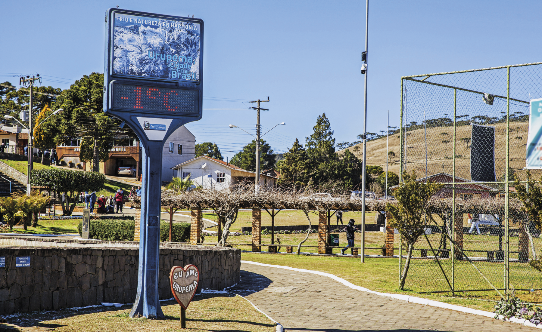 Fotografia. Termômetro de rua em uma praça registrando menos 1 grau Celsius. Ao fundo, é possível identificar casas, árvores, chão gramado e pessoas agasalhadas caminhando. O céu da paisagem exibida na imagem está azul e sem nuvens.