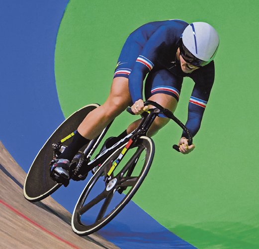 Fotografia: Atleta mulher está pedalando bicicleta em uma pista profissional azul e verde. A atleta veste um macacão azul, um capacete cinza e óculos escuros.