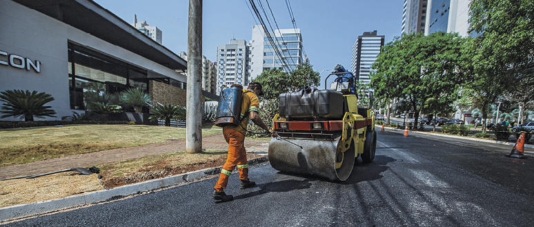 Fotografia: Um homem, com roupa laranja, está utilizando uma máquina para asfaltar o trecho de uma rua. O dia está ensolarado, ao fundo da imagem, prédios e árvores.