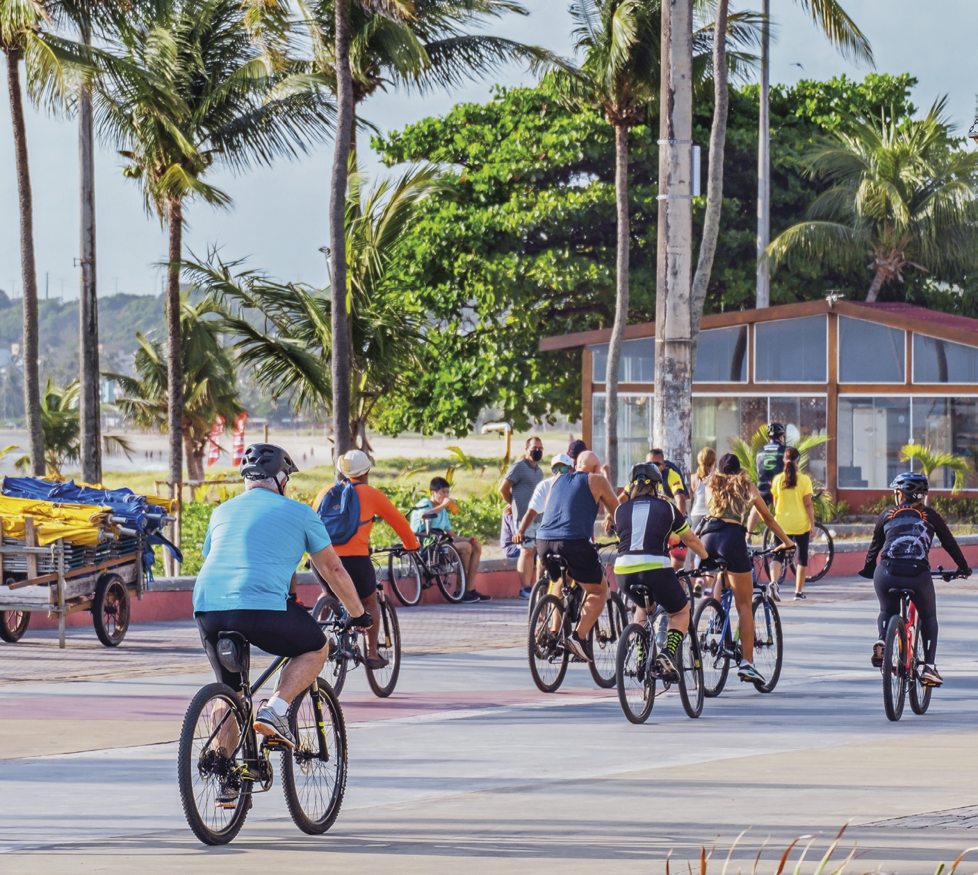 Fotografia. Calçadão à beira mar, com 6 pessoas andando de bicicleta, duas pessoas com bicicleta paradas e duas pessoas caminhando Há também um carrinho para transporte de guarda sóis. Do lado esquerdo do calçadão há um gramado com palmeiras. À esquerda do calçadão, mas à direita da imagem, um quiosque vermelho envidraçado. Mais ao fundo, areia seguida de morros e um céu azul acinzentado.