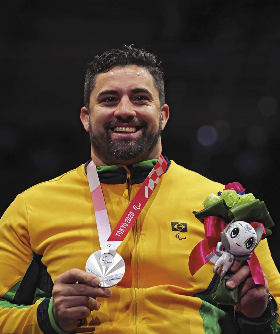 Fotografia. Homem branco, sorridente exibindo com a mão direita a medalha de prata conquistada. Com a mão esquerda, ele segura um boneco que representa o mascote dos jogos paralímpicos de Tóquio. Ele utilizada agasalho da seleção brasileira.
