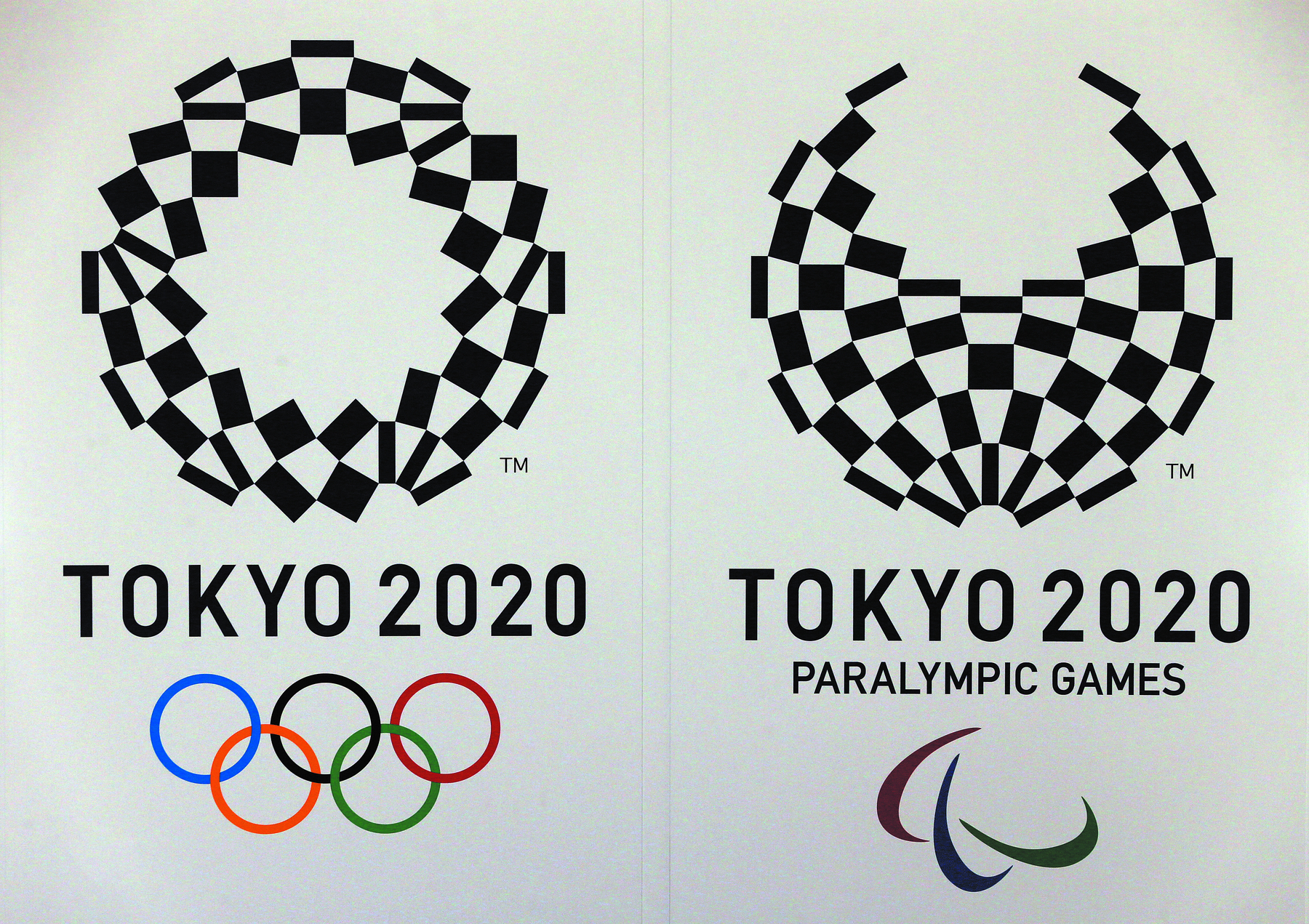 Fotografia. 2 símbolos dos jogos olímpicos e paralímpicos. À direita, na parte superior, há uma figura de um símbolo, composta por vários retângulos em preto que um globo. Abaixo do globo está escrito TOKYO 2020. Na parte inferior, há seis argolas coloridas entrelaçadas. 

À esquerda, na parte superior, há uma figura de um símbolo, composta por vários retângulos em preto que se unem para formar um globo, sendo a parte superior é aberta acima, abaixo escrito TOKYO 2020 paralympic games. Na parte inferior, há três linhas curvas nas cores vermelha, azul e verde.