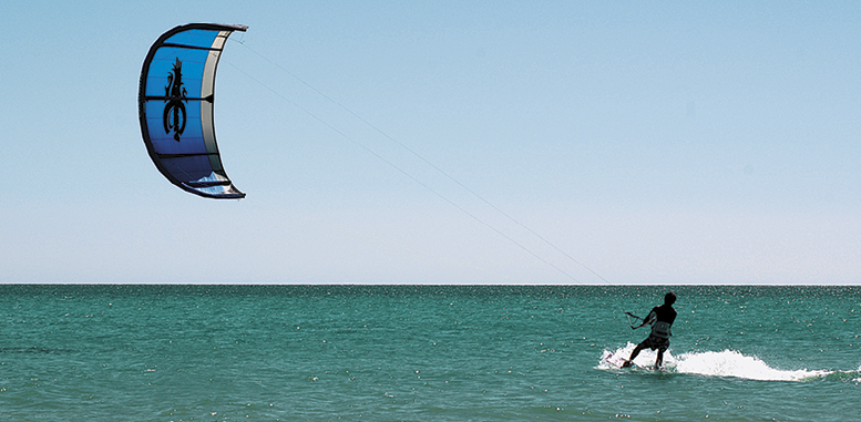 Fotografia. Vista frontal de um homem no mar segurando no ar uma pipa grande em formato de arco.