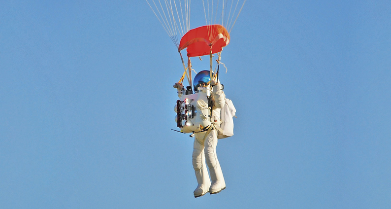 Fotografia: Um astronauta, trajado com roupa própria branca, óculos azul, carregando uma mochila com cilindro de oxigênio nas costas e outra no tórax, está pousando, com um paraquedas vermelho aberto. Ao fundo um céu azulado, sem nuvens.