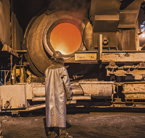 Fotografia: Um homem com uma roupa especial cinza está de costas olhando para uma máquina de fundição de alumínio.
