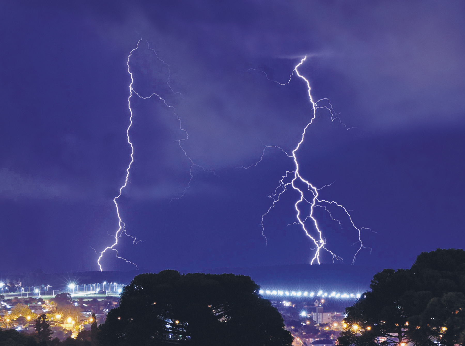 Fotografia. Vista noturna de uma cidade em uma noite com raios. Dois raios descem do céu. Algumas árvores estão próximas.