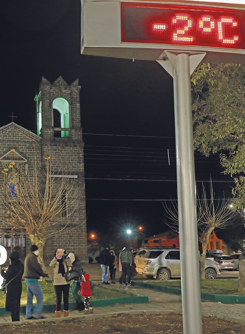 Fotografia. Imagem de um termômetro em frente a uma igreja, algumas pessoas agasalhadas e no termômetro marca menos 2 graus célcios.