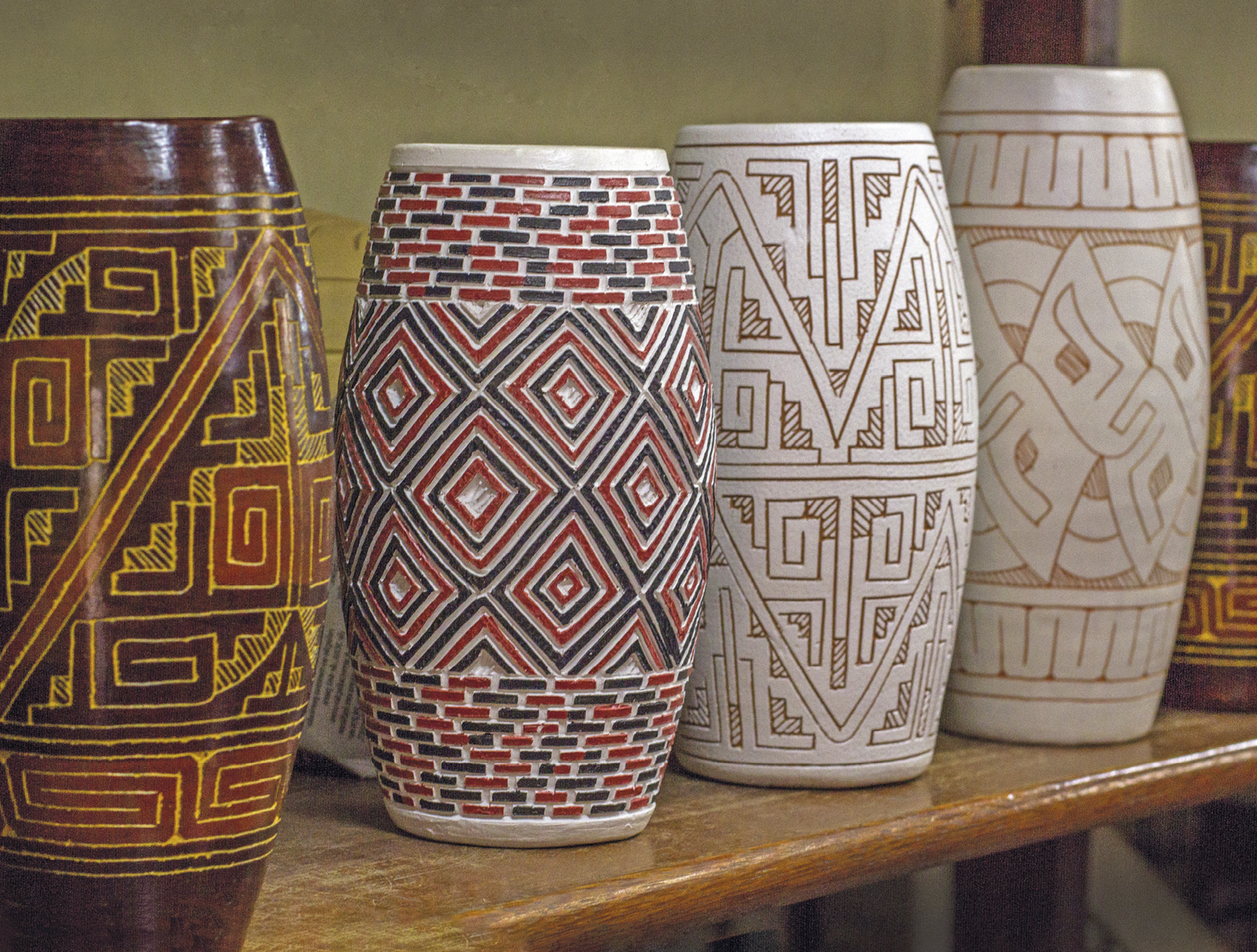 Fotografia. Vasos cilíndricos de cerâmicas. Da esquerda para direita: vaso marrom com formas geométricas douradas e avermelhadas. Vaso branco com formas geométricas em formato de losangos e retângulos na cor vermelha e preta. Dois vasos de cor clara com padrões geométricos na cor marrom.