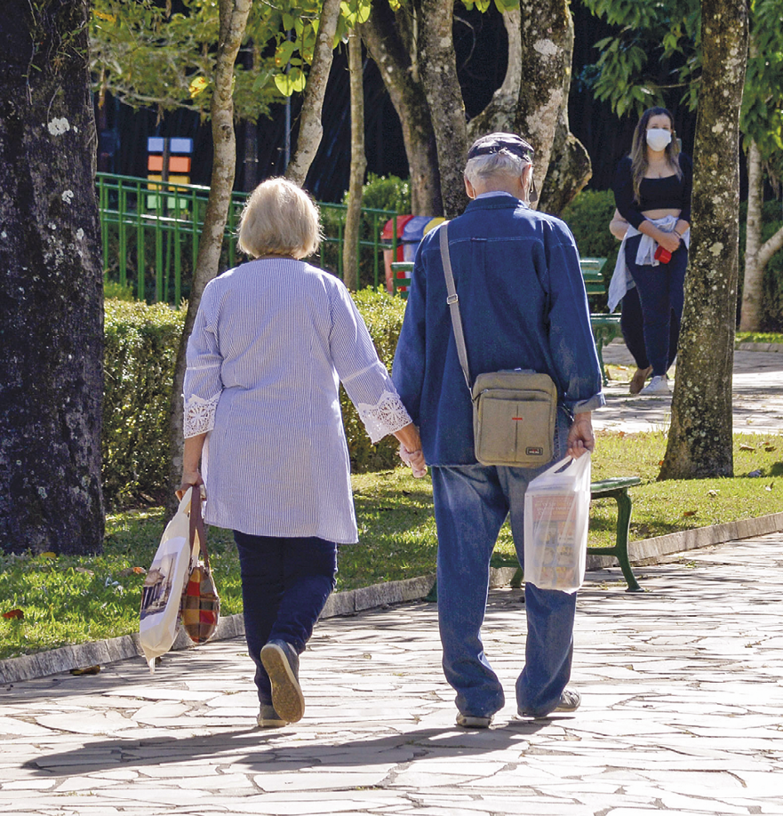 Fotografia vista de trás de um casal de idosos caminhando de mãos dadas em um parque. À esquerda, grama e árvores.