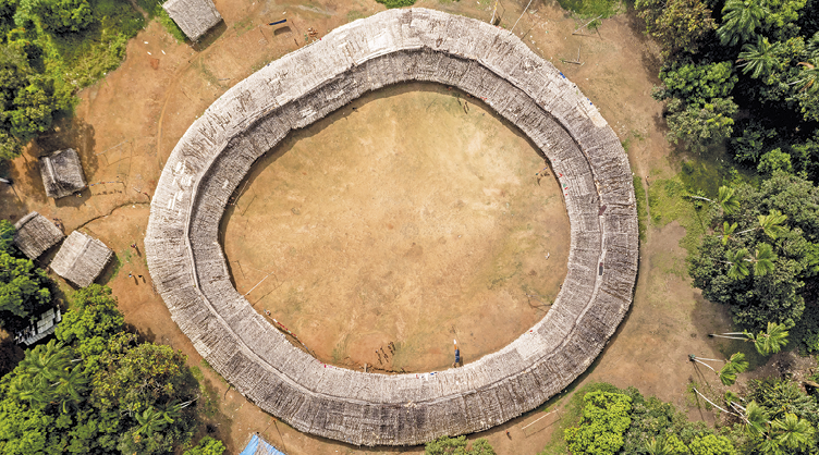 Fotografia. Vista aérea de uma aldeia circular em solo marrom. Nas laterais, vegetação.