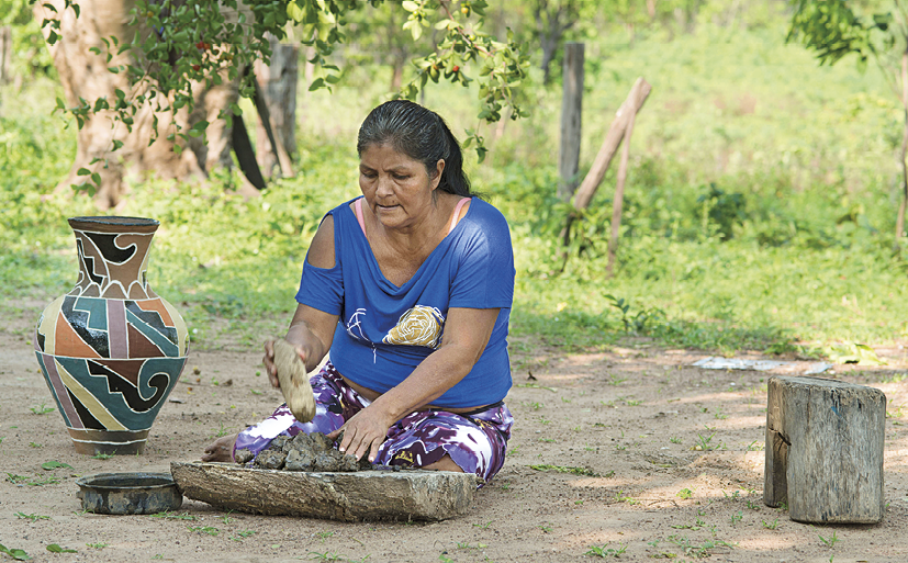 Fotografia. Mulher indígena de cabelo preto e blusa azul. Ela está sentada no chão com um objeto nas mãos. Ao lado, vaso de cerâmica colorido. Ao fundo, vegetação.