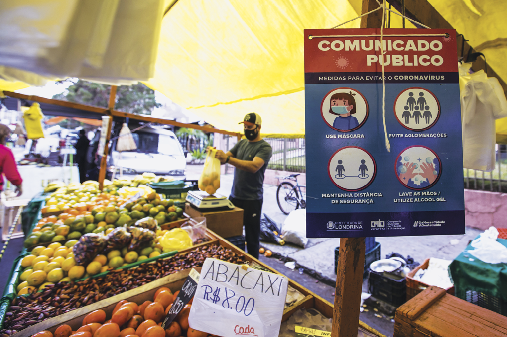 Fotografia. Parte de uma feira livre com barracas com frutas. Abacaxi: 8 reais. Ao lado, placa escrita: COMUNICADO PÚBLICO com a imagem de uma pessoa com máscara de proteção cobrindo a boca e o nariz. Grupo de pessoas, duas pessoas afastadas e duas mãos com água e sabão.