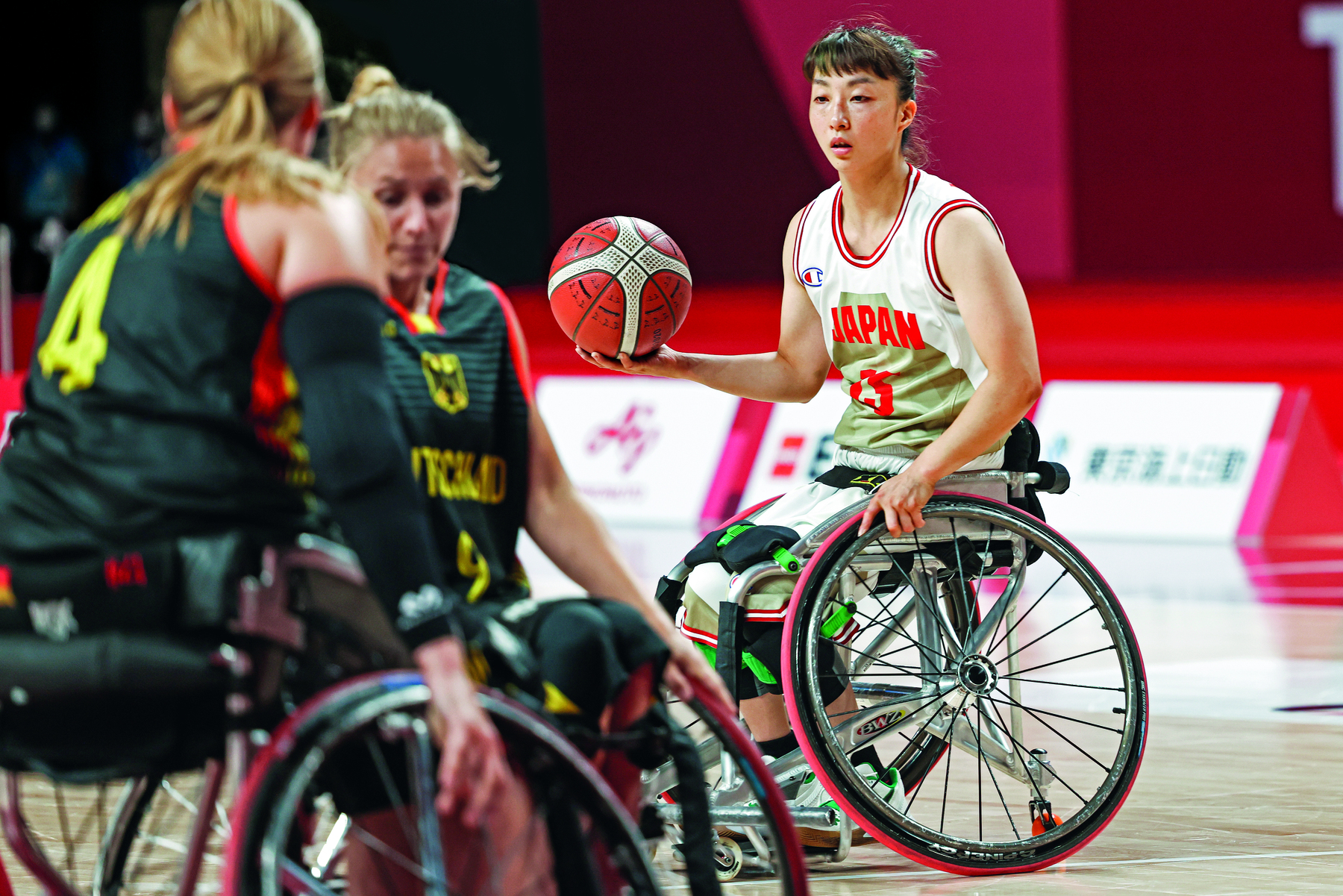 Fotografia. Atletas cadeirantes das equipes femininas de Alemanha e Japão disputando uma partida de basquete. A bola está nas mãos de uma das atletas do Japão.
