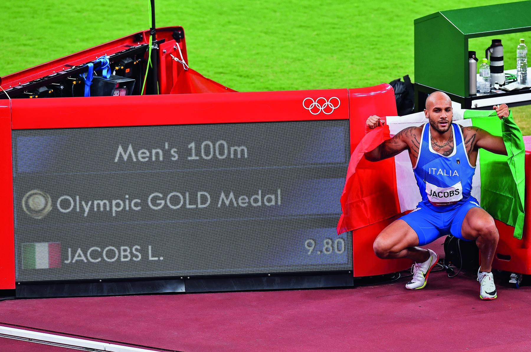 Fotografia. Homem negro de regata azul e bermuda está abaixado com os braços para cima. À esquerda, quadro: Men’s 100 m. Olympic GOLD Medal. Abaixo, bandeira da Itália e o nome JACOBS L.