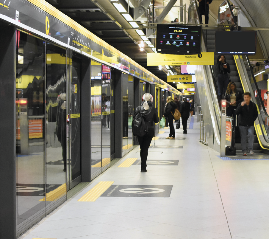 Fotografia. Vista da plataforma do metrô com algumas pessoas. Há portas de vidros antes do embarque e desembarque. À direita, escadas rolantes