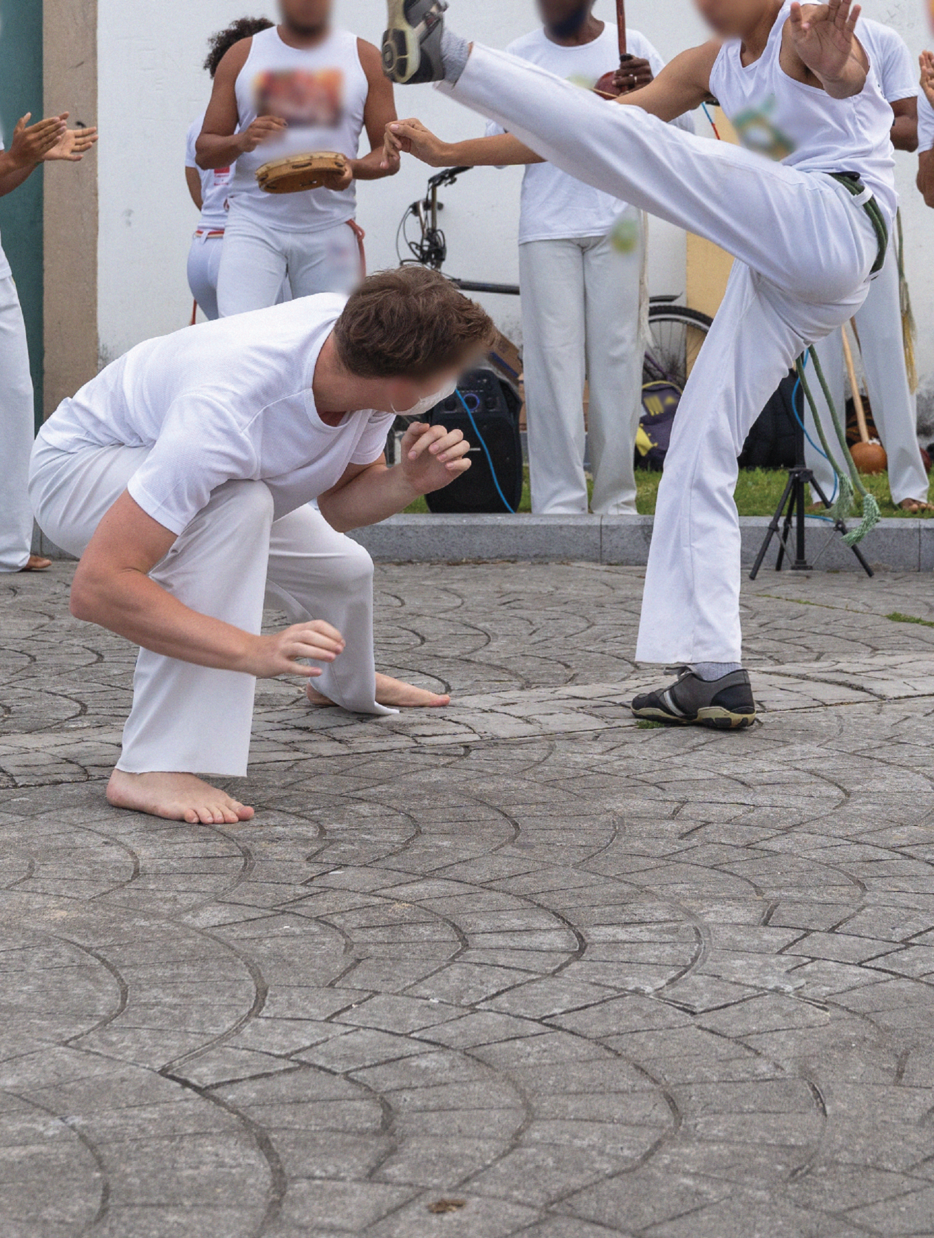 Fotografia. Grupo de pessoas de camisetas e calças brancas jogando capoeira em uma rua.
