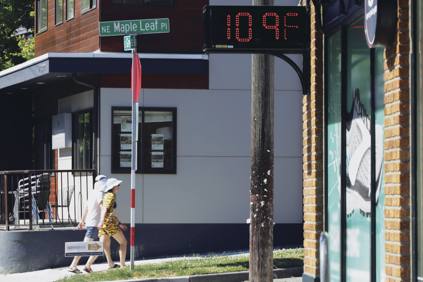 Fotografia. Termômetro de rua registrando 109 graus fahrenheit. O dia está ensolarado e há duas pessoas caminhando na rua, utilizando roupas leves e chapéu.