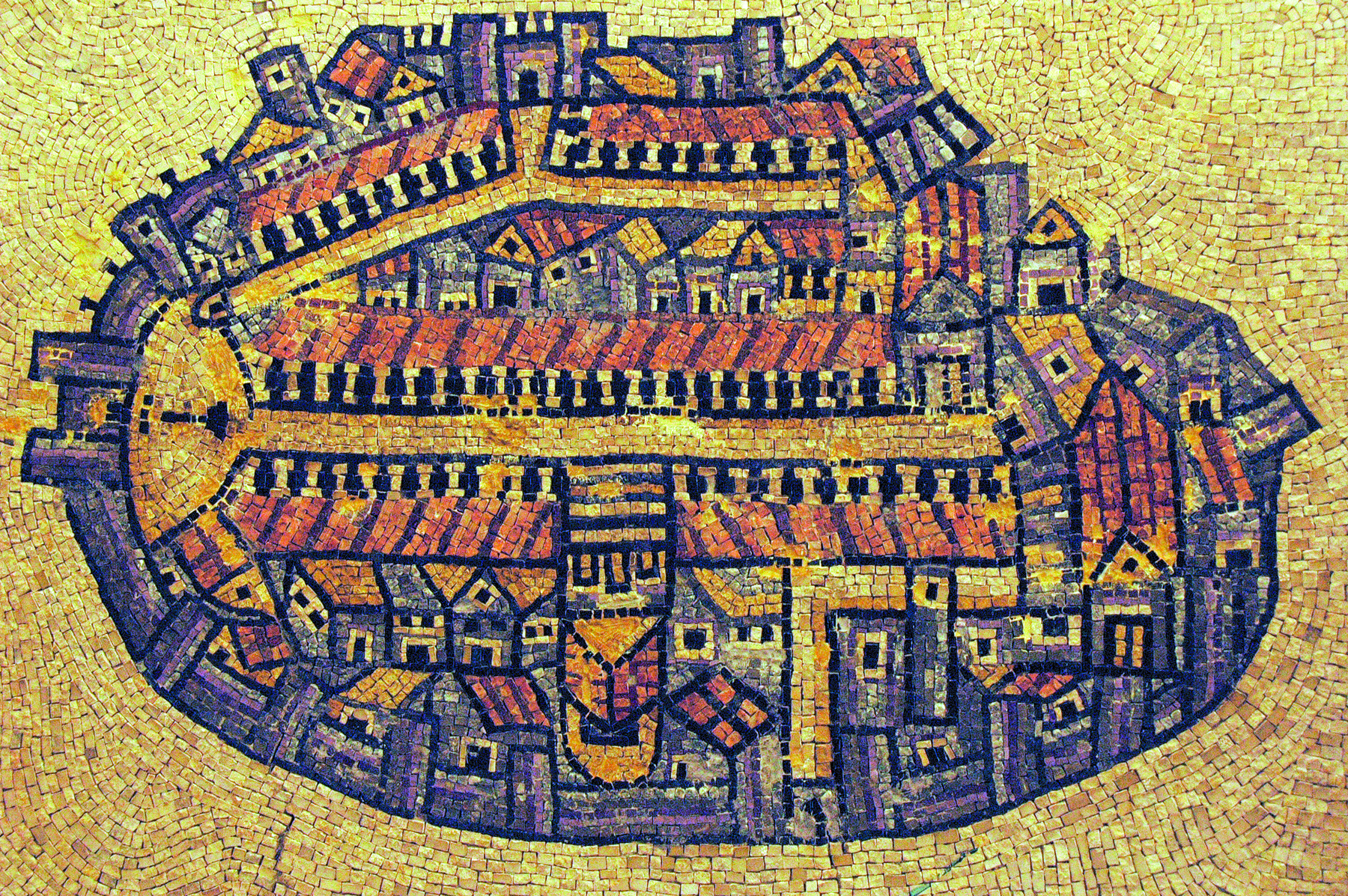 Fotografia. Reprodução em mosaico de pedras do mapa de uma cidade. O formato é semelhante ao de um círculo achatado e o fundo é amarelado. É possível observar uma espécie de entrada, à esquerda, uma rua central horizontal e telhados e casas.