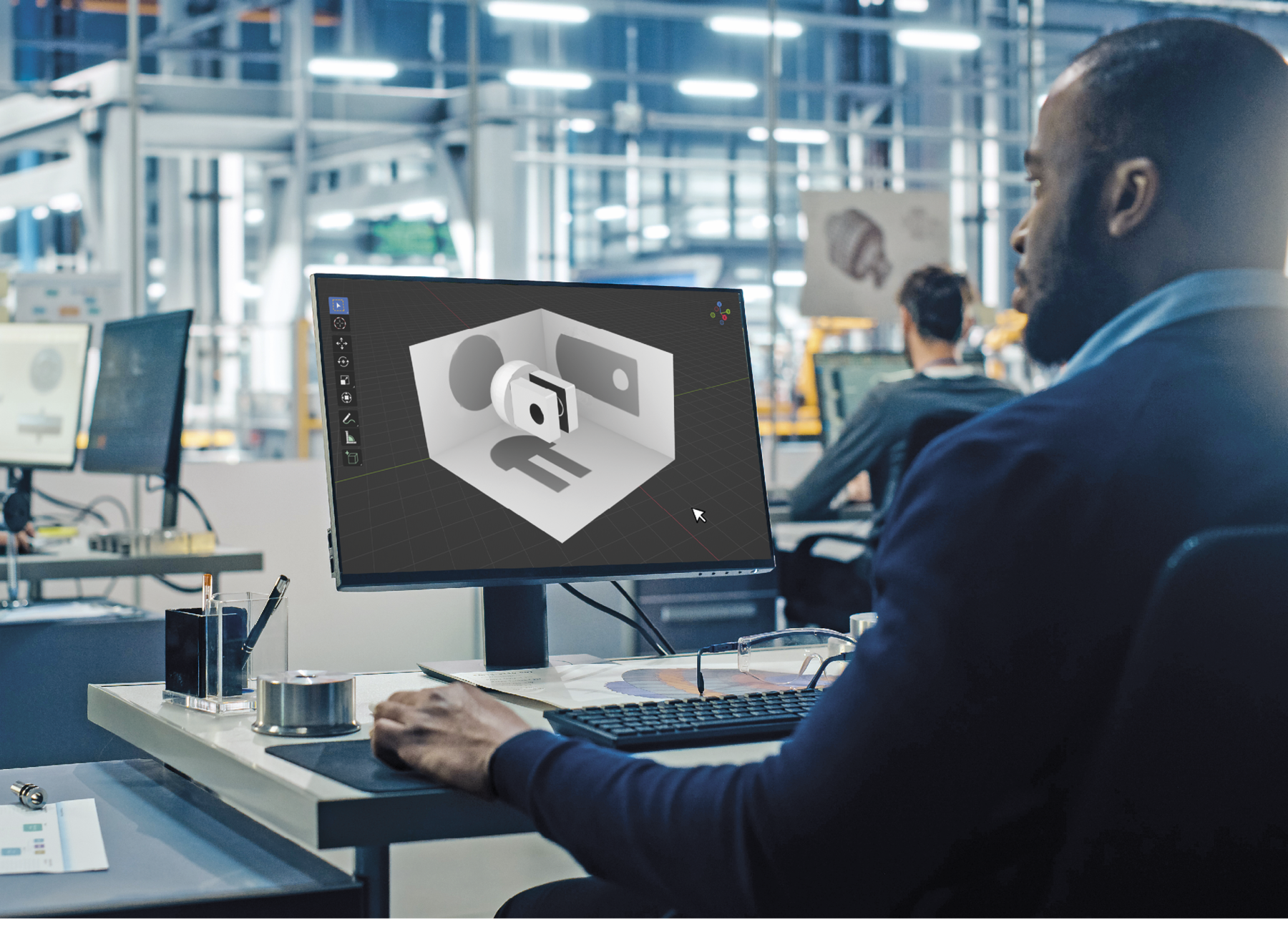 Fotografia. Vista de trás de um homem negro de cabelo curto e blusa azul. Ele está de frente para um monitor com a imagem de uma peça em 3D com as projeções em representadas em planos.