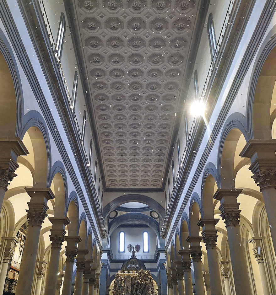 Fotografia. Vista de baixo para cima do do interior da igreja representada no desenho anterior. É possível observar pilares do lado direito e do lado esquerdo que formam arcos na parte superior. Uma parte do teto aparece bem nítida com desenhos em formato circular.