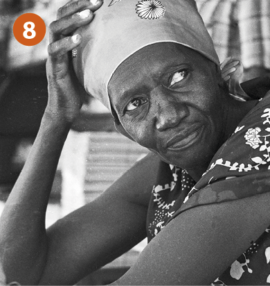 Fotografia 8. Fotografia em preto e branco de mulher negra de lenço claro na cabeça e camiseta regata. Ela está com um dos braços apoiado na cabeça.