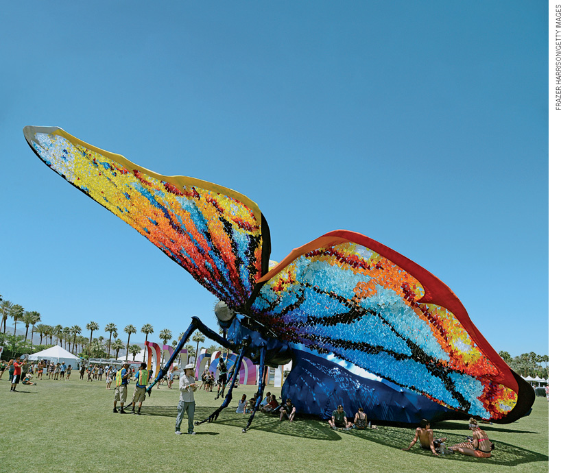 IMAGEM: escultura de grandes dimensões de um inseto com asas coloridas, semelhante a uma borboleta. a instalação está em um gramado, a céu aberto, e pessoas estão sentadas à sua sombra ou a apreciando. FIM DA IMAGEM.