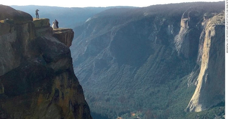 IMAGEM: um casal de noivos está no alto de uma montanha posando para uma fotografia. no fundo, há uma parede de pedra com vegetação. FIM DA IMAGEM.