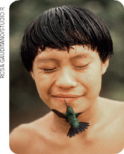IMAGEM: um menino indígena sorri. um beija flor voa com o bico tocando em sua boca. FIM DA IMAGEM.