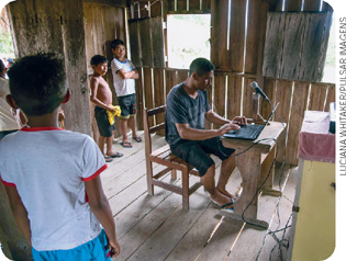 IMAGEM: em uma casa com cão e paredes de madeira, três meninos observam um homem operar um computador com um microfone conectado a ele. FIM DA IMAGEM.