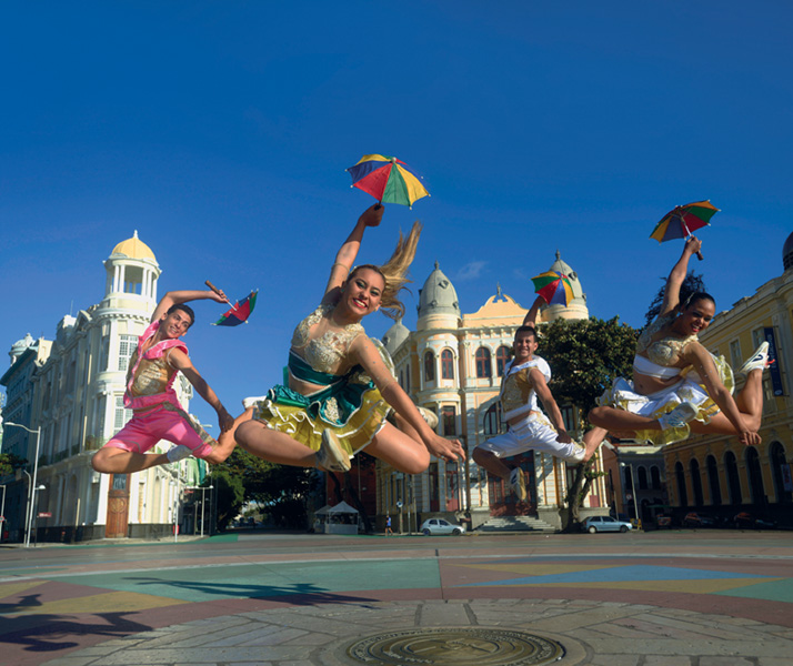 IMAGEM: no início da unidade seis, uma foto apresenta quatro dançarinos de frevo em uma praça. são duas mulheres e dois homens que usam roupas tradicionais de frevo e seguram uma sombrinha. eles pulam alto e sorriem. FIM DA IMAGEM.