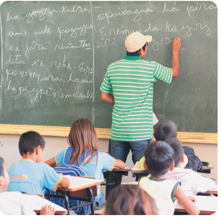IMAGEM: uma sala de aula de uma escola indígena, em que as crianças estão em carteiras e o professor escreve na lousa. O professor escreve em guarani. FIM DA IMAGEM.