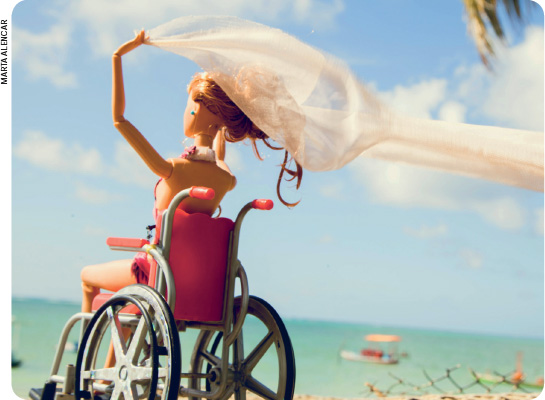 IMAGEM: uma boneca em cadeira de rodas está em uma praia, segurando um véu contra o vento. ela ergue os braços e segura o véu acima da cabeça. FIM DA IMAGEM.