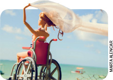 IMAGEM: uma boneca em cadeira de rodas está em uma praia, segurando um véu contra o vento. ela ergue os braços e segura o véu acima da cabeça. FIM DA IMAGEM.