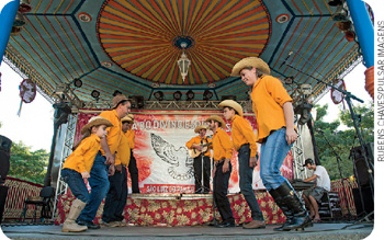 IMAGEM: um grupo formado por adultos e crianças dança em um coreto. eles usam botas e chapéus de vaqueiro e formam duas filas. no fundo, em um palco, há homens que cantam e tocam violão. FIM DA IMAGEM.
