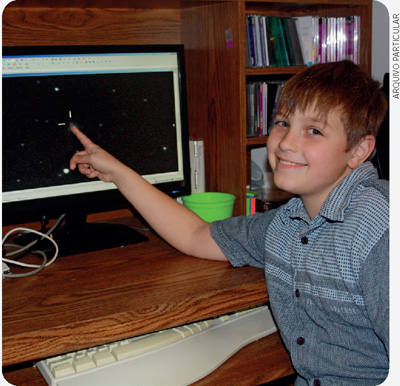 IMAGEM: fotografia de nathan gray, um menino sorridente sentado em frente a um computador. ele aponta para a tela do computador com o dedo indicador. FIM DA IMAGEM.