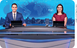 IMAGEM: um homem e uma mulher sentados atrás de uma bancada em um cenário de um telejornal. na frente deles, sobre a bancada, há computadores. FIM DA IMAGEM.
