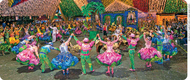 IMAGEM: pessoas com trajes típicos de dança de quadrilha formam uma roda em uma festa junina. o espaço está enfeitado com bandeiras penduradas. FIM DA IMAGEM.