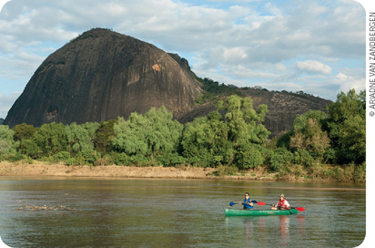 IMAGEM: duas pessoas remam em uma canoa sobre o rio. no fundo, há vegetação densa e montanhas de pedra. FIM DA IMAGEM.
