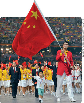 IMAGEM: pessoas comemoram com pequenas bandeiras da china no encerramento dos jogos olímpicos de pequim. na frente, o menino xiao li e o atleta yao ming. o atleta segura uma haste comprida com a bandeira do país. FIM DA IMAGEM.
