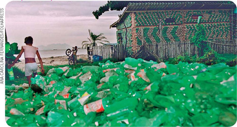 IMAGEM: pilha de garrafas de plástico recolhidas em uma praia. no fundo, uma casa construída com partes de garrafas plásticas e outros materiais. FIM DA IMAGEM.