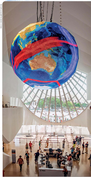 IMAGEM: grande esfera de led suspensa no interior de um museu, nela, a imagem da terra. abaixo, o museu está cheio de visitantes. FIM DA IMAGEM.