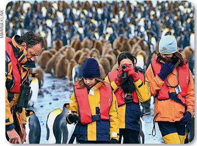 IMAGEM: amyr e as filhas na antártica. uma das meninas tira fotos e todos usam coletes salva-vidas. no fundo, centenas de pinguins sobre o gelo. FIM DA IMAGEM.
