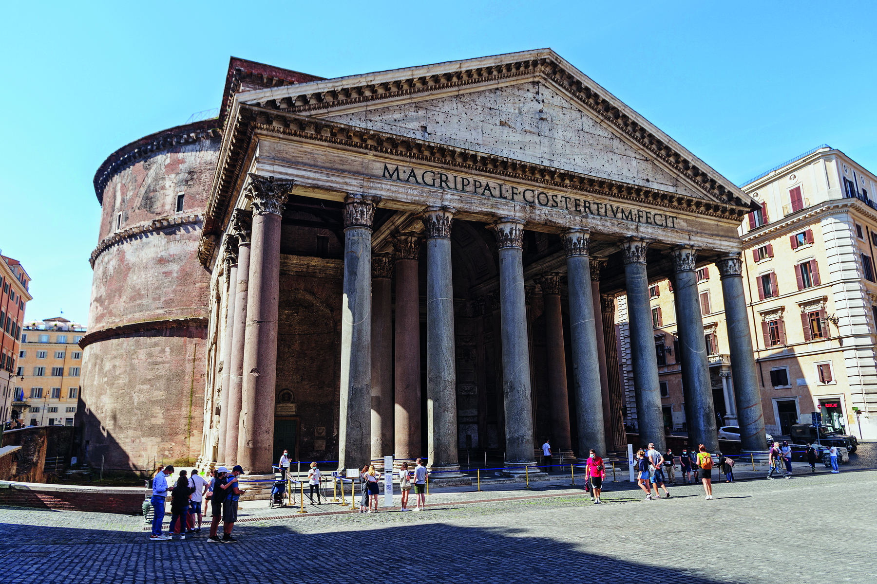 Fotografia. Vista da fachada de um panteão, com colunas frontais, um teto triangular e escritas no topo, na horizontal. À frente, pessoas transitam.