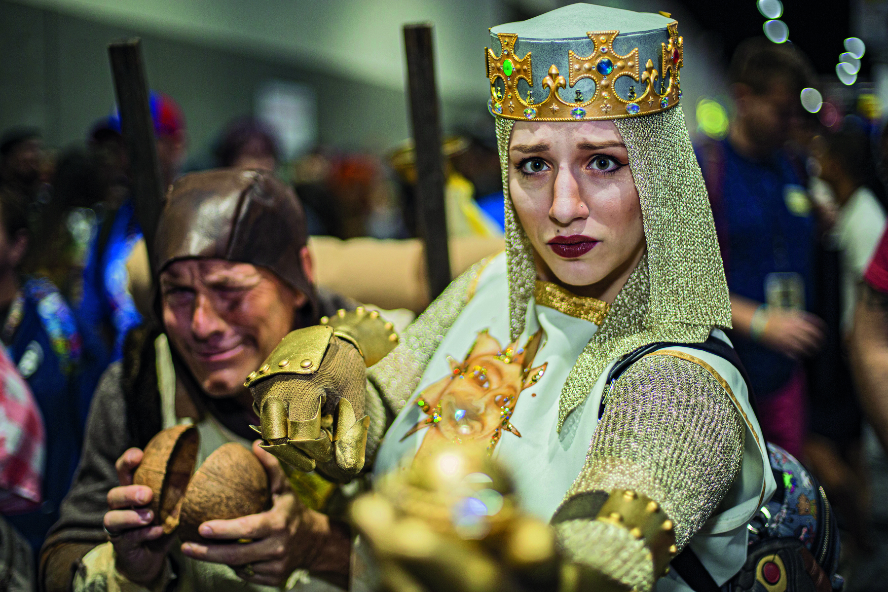 Fotografia. Destaque para uma mulher vestindo uma fantasia de armadura, com luvas metálicas, uma coroa dourada na cabeça com pedras verdes e azuis, e redes de tecidos caindo na lateral de seu rosto.