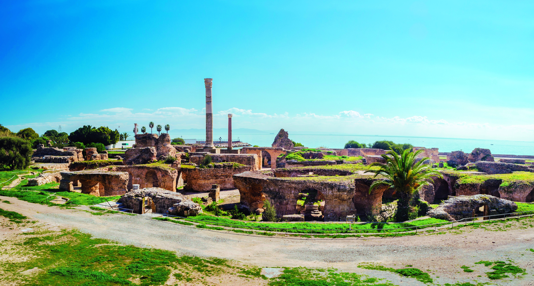 Fotografia. Vista de ruínas de uma cidade à beira mar, com construções em formato circular, corredores e duas colunas verticais ao fundo.
