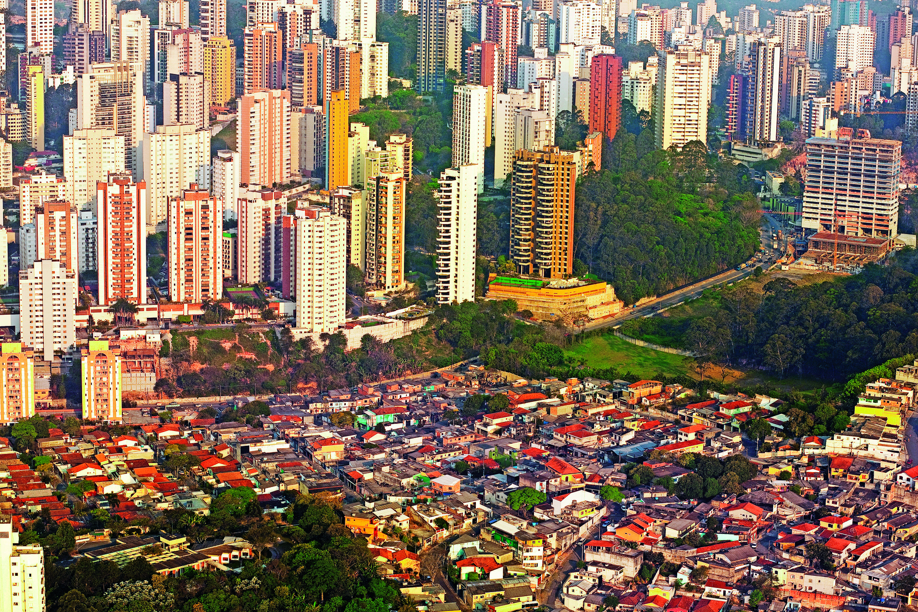 Fotografia. Vista aérea de uma cidade com dois agrupamentos de moradias distintas. Em primeiro plano há diversos casebres muito próximos. Ao fundo há diversos prédios grandiosos.