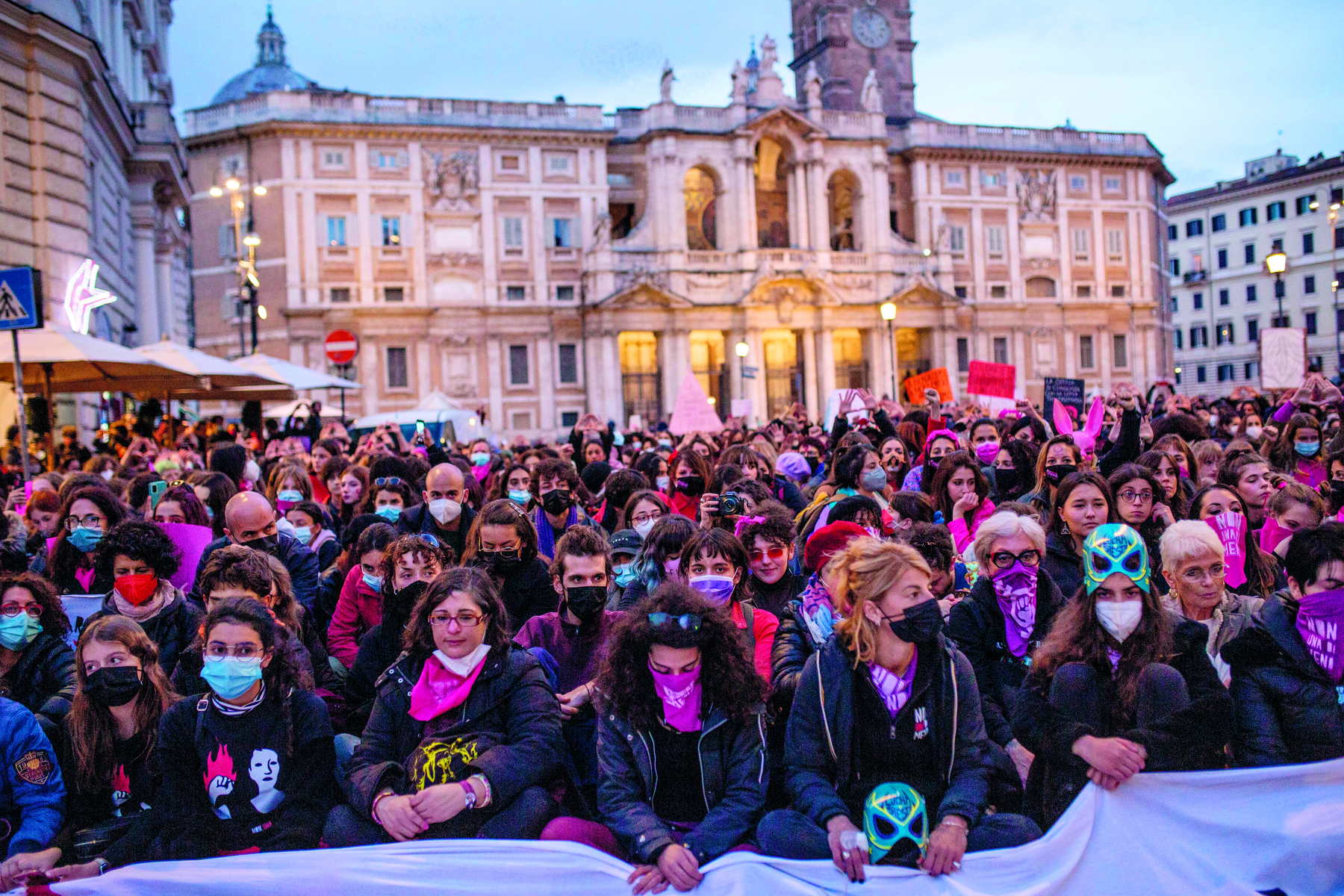 Fotografia. Uma multidão de mulheres de diferentes idades, lado a lado, segurando faixas. Muitas delas usam máscaras e bandanas cobrindo partes de seus rostos.