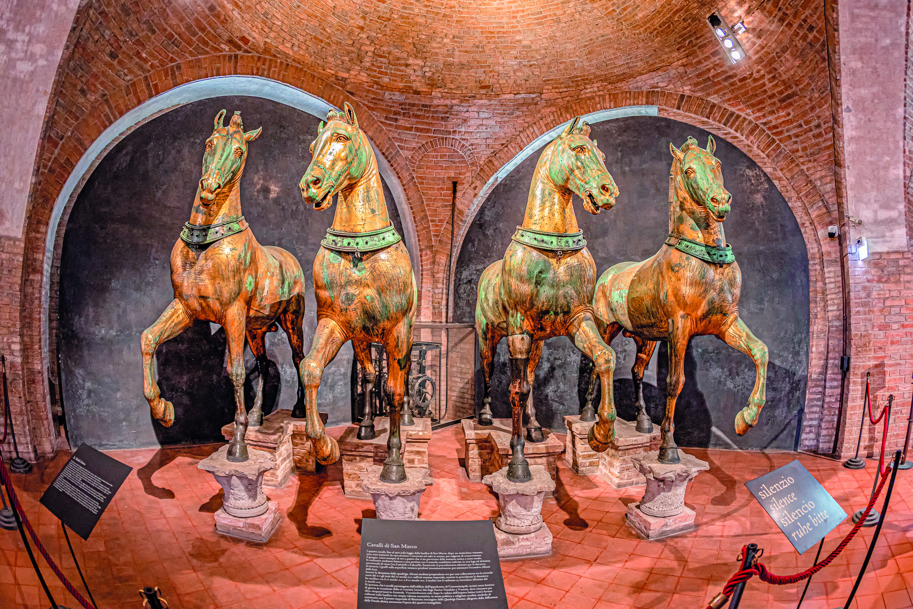 Esculturas. Quatro cavalos metálicos em exposição. Estão lado a lado, com as patas em suportes no chão. Ao fundo, paredes de tijolos.