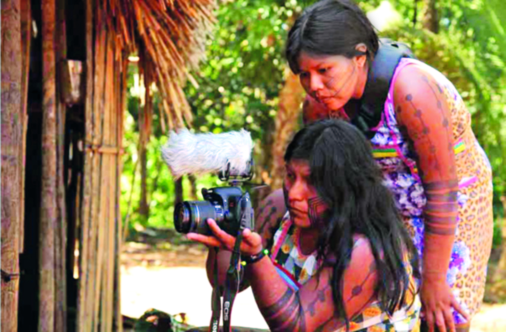 Fotografia. Destaque para duas mulheres indígenas, uma delas em posição mais baixa, segurando uma máquina fotográfica com as mãos. A outra, por detrás, tem o olhar voltado para o objeto. Ambas têm longos cabelos lisos e escuros e usam vestidos coloridos e pinturas corporais